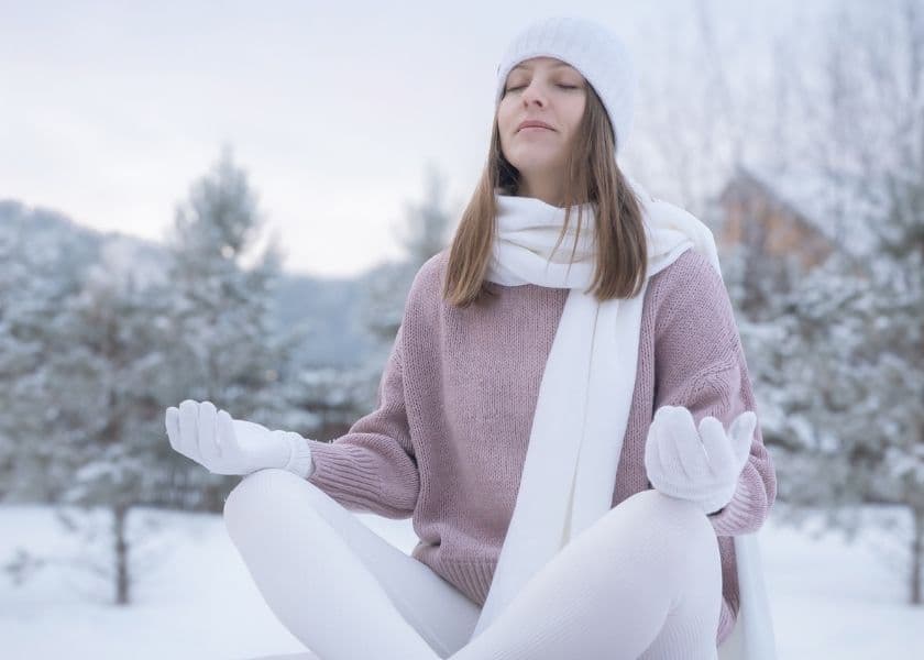 a meditating woman sitting cross-legged in a sweater, hat, and gloves outside with a snowy, tree-lined backdrop a meditating woman sitting cross-legged in a sweater, hat, and gloves outside with a snowy, tree-lined backdrop