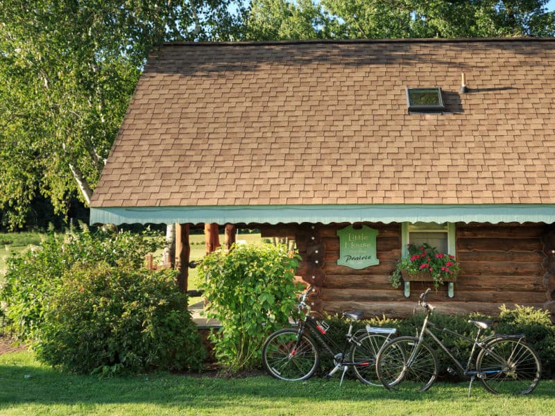 Exterior of log cabin with two bicycles standing under a window with a windowbox filled with blooming flowers, and an open field in background.