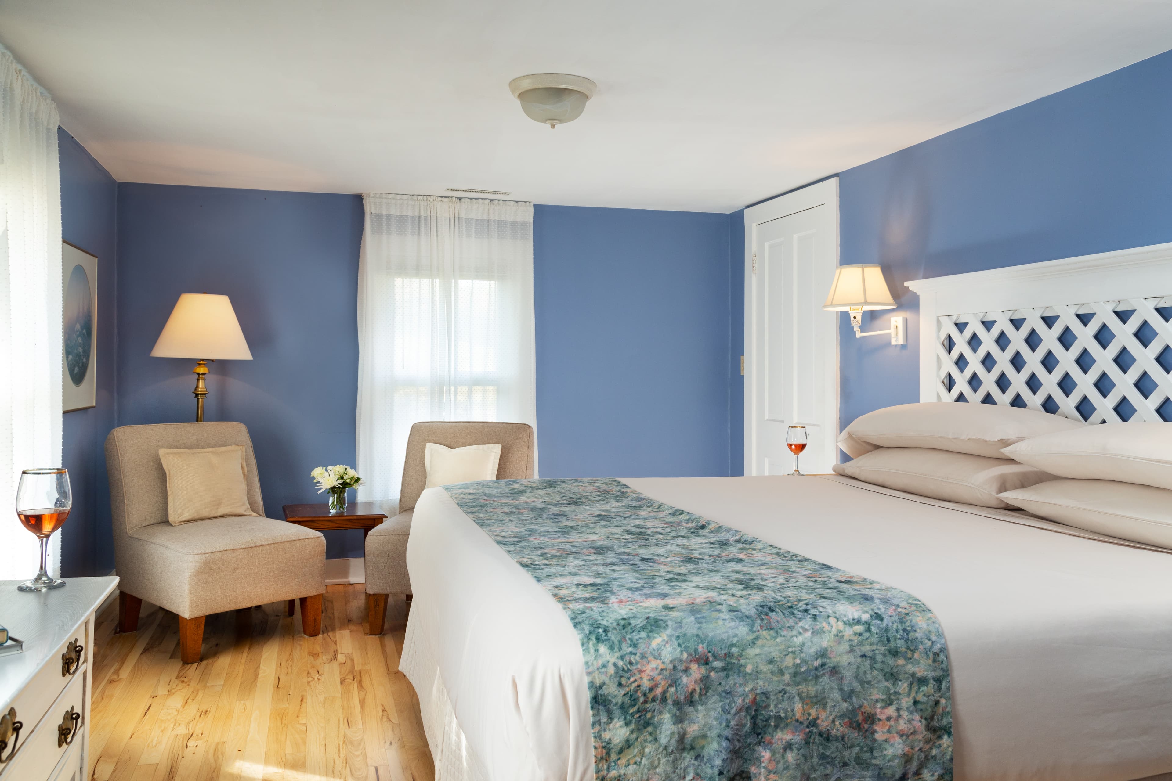 King bed with white lattice headboard, wall mounted reading lights on both sides of bed, sitting area with two upholstered chairs near light filled window.