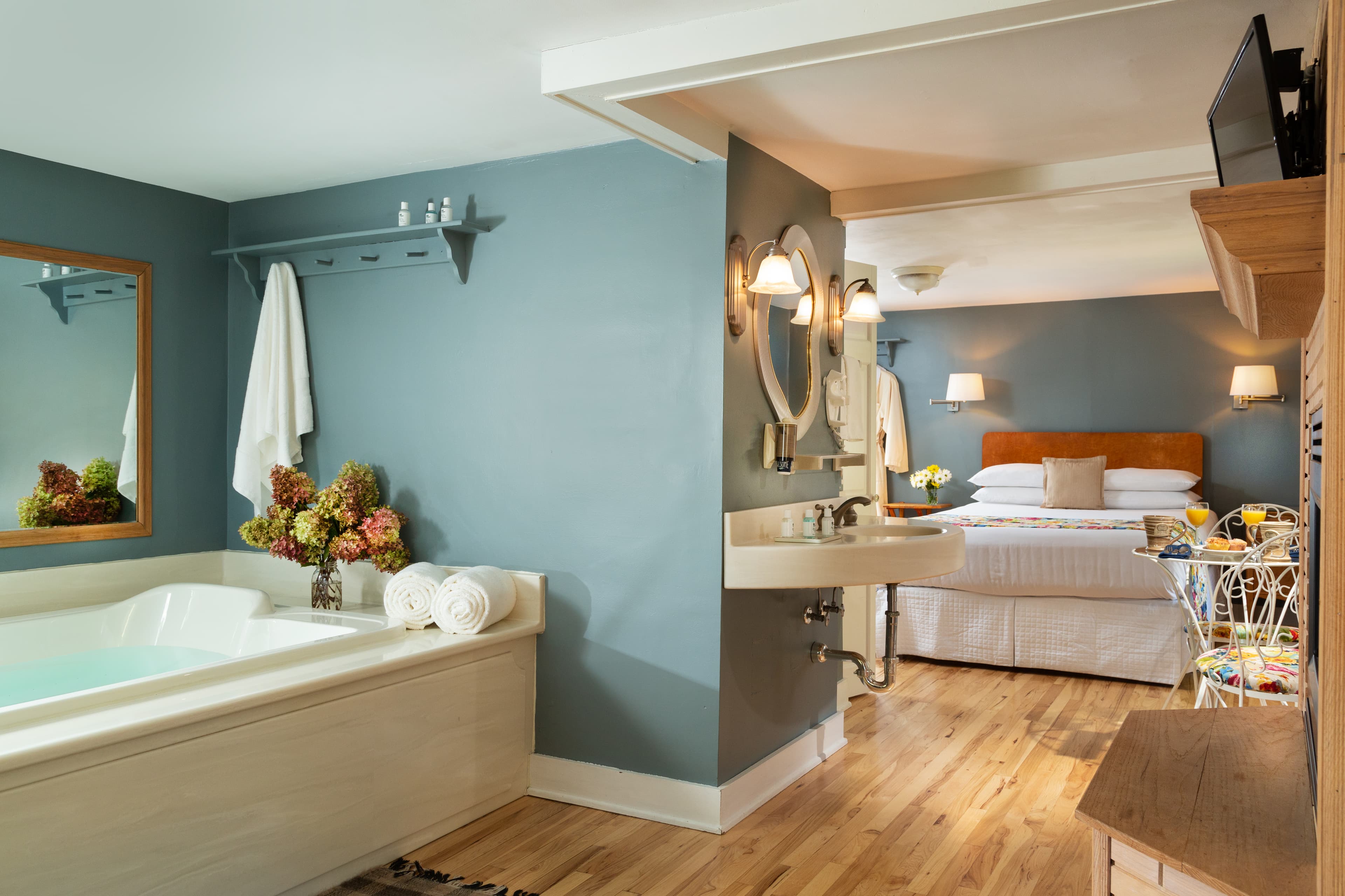 Tranquil suite with double Bain Ultra whirlpool tub to the left in foreground separated with partial wall from sleeping area with queen bed and wall mounted reading lamps on either side.