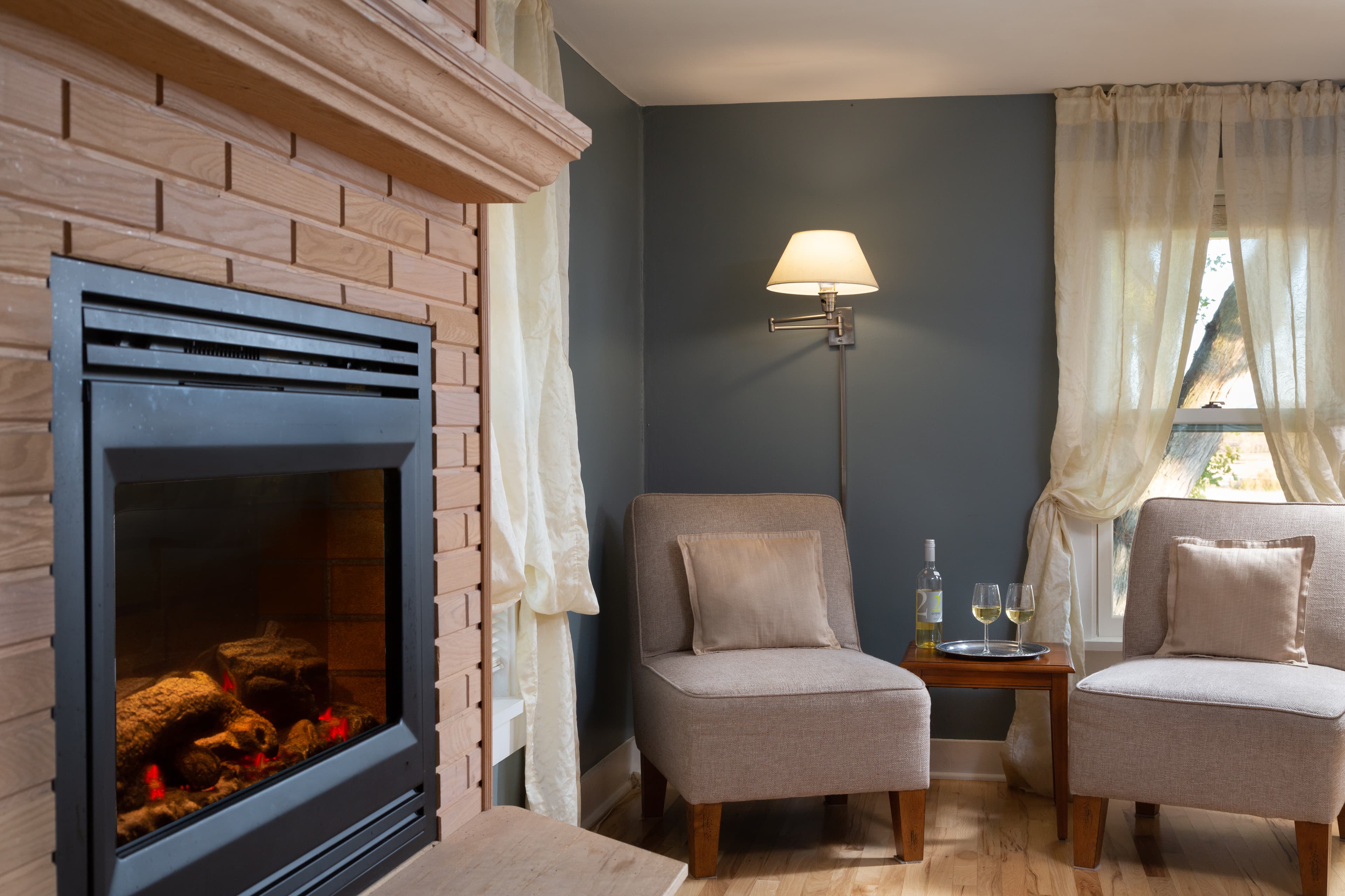 Sitting area with oak wood brick surround on fireplace, two upholstered chairs, little table, and a wall mounted lamp.