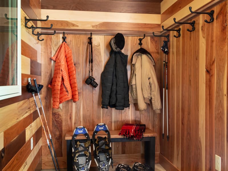 Mud room with natural wood walls and large black hooks some empty and some hung with winter coats, a bench sits on the floor, and there are ski boots and ski poles leaning against the wall.