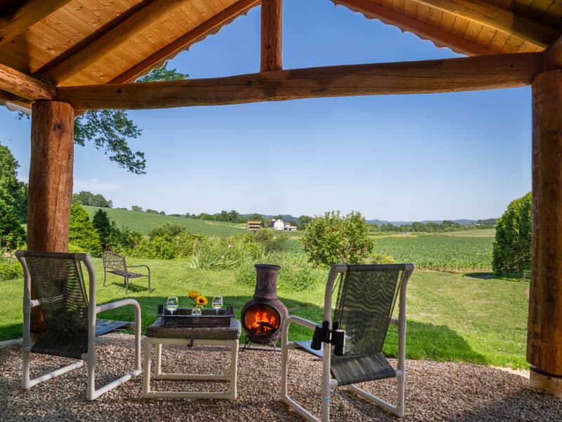 Outdoor patio area with log roof overhead, two chairs with a small table and a chiminea with a fire glowing inside. There is a beautiful pastoral view of open green fields and a cloudless blue sky.
