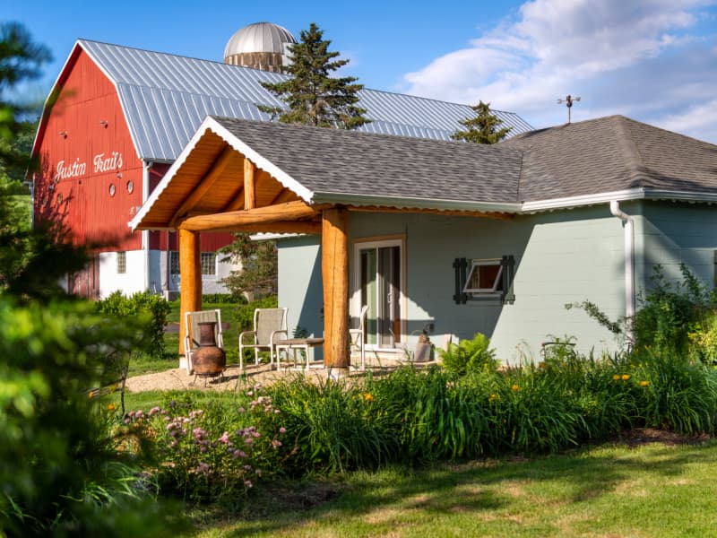 Exterior of the Sunrise Cottage with covered patio and shrubbery and a big red barn with steel roof just beyond.