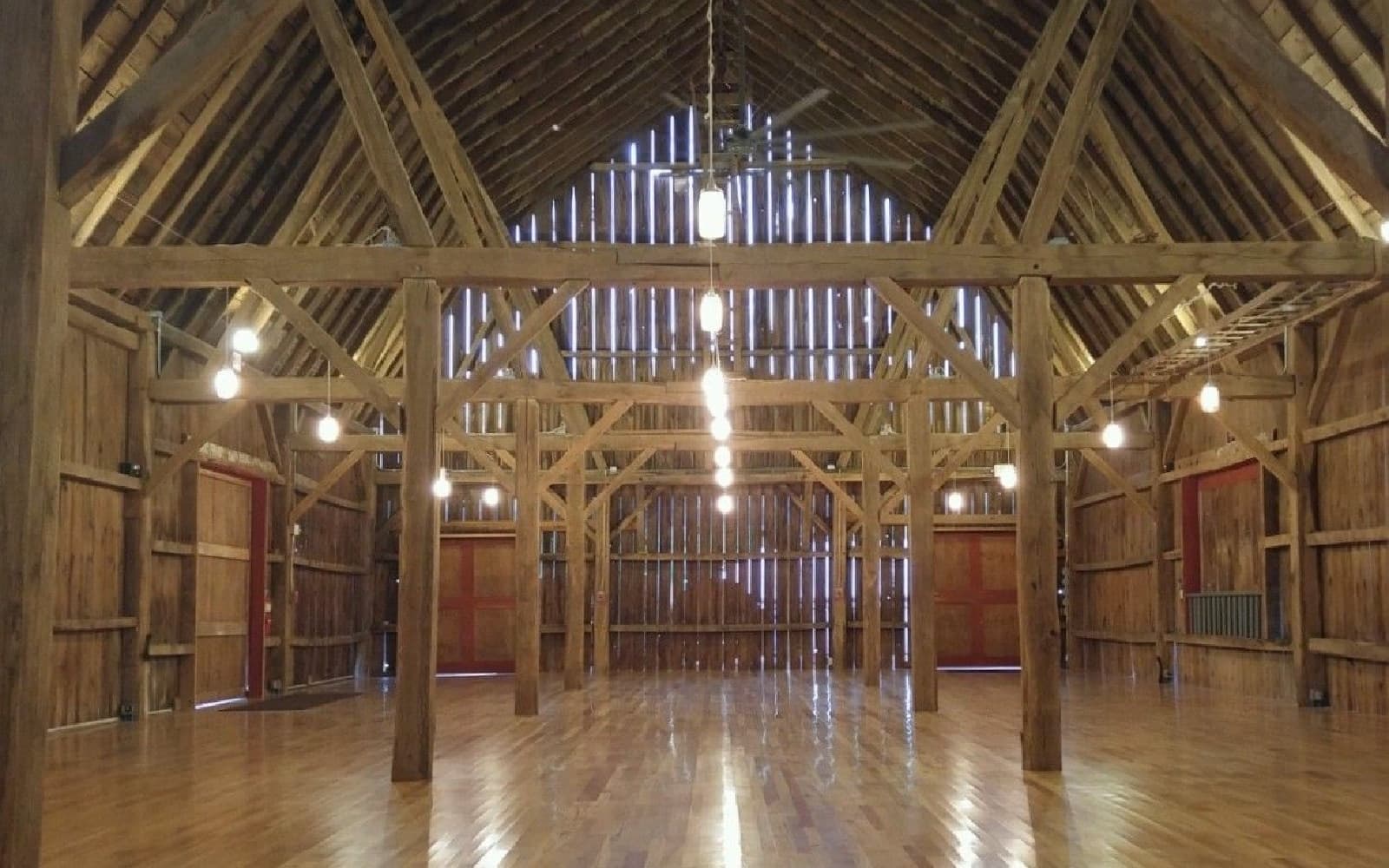 Interior of a spacious wooden barn with high ceilings and wooden beams, featuring pendant lights and polished wooden floors.