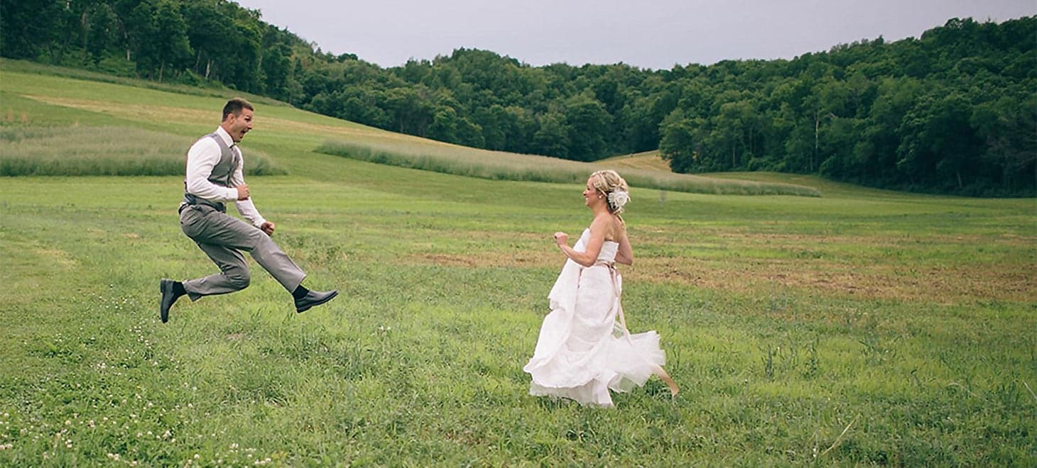 A groom joyfully leaps toward a bride in a lush, green field.