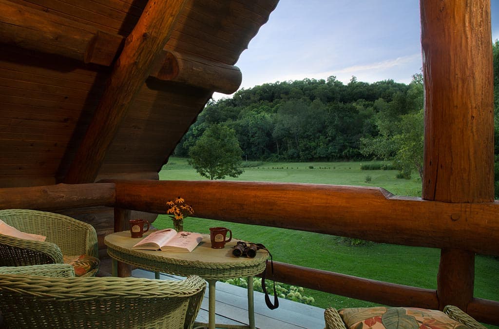 A cozy log cabin balcony with wicker chairs overlooks a lush green field at dusk.