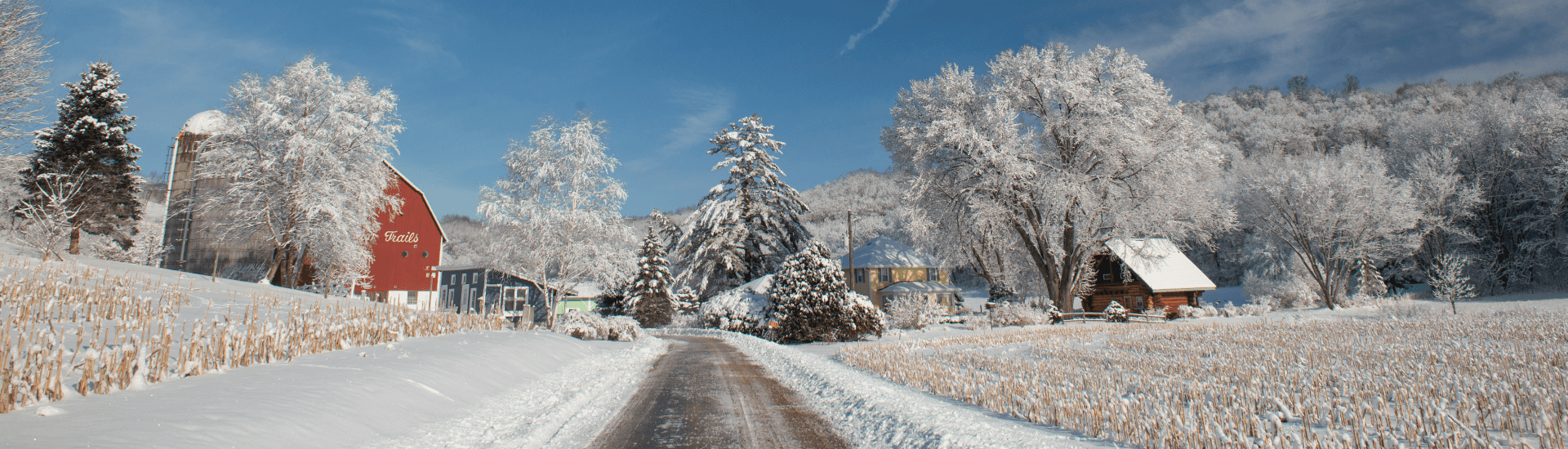 A picturesque winter farm scene featuring barns, trees, and a snow-covered road.
