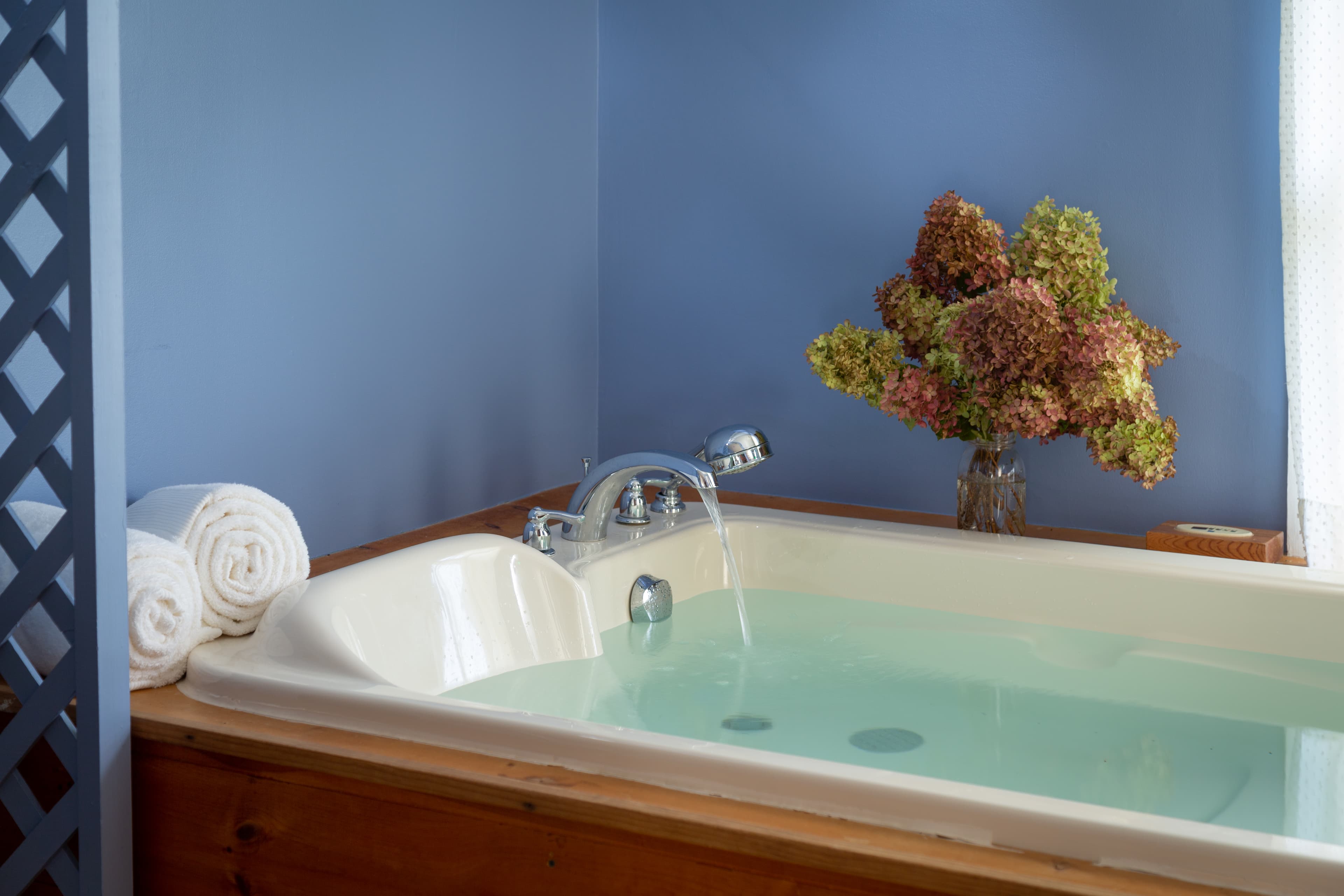 Large double BainUltra whirlpool bathtub with natural wood surround and plush white towels rolled up on the ledge.