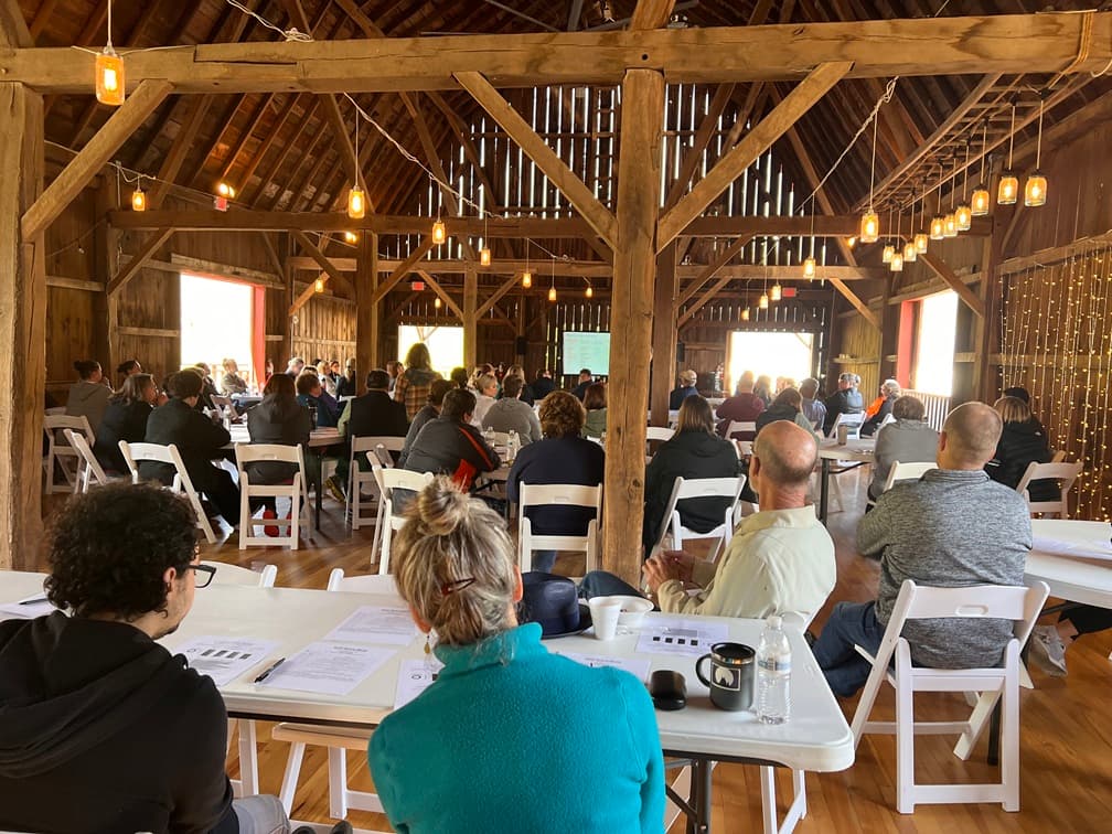 A group of attendees sits at tables in a rustic barn, listening to a presentation.
