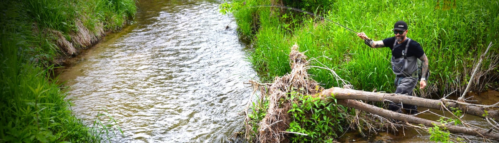 A fisherman stands by a flowing creek, casting a line into the water amidst lush greenery.